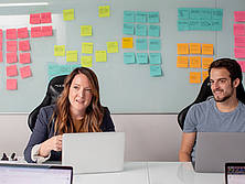 Zwei Kollegen, eine Frau und ein Mann sitzen mit ihren Laptops an einem weißen Tisch. Hinter ihnen kleben bunte Post-it-Notizzettel an einer weißen Board